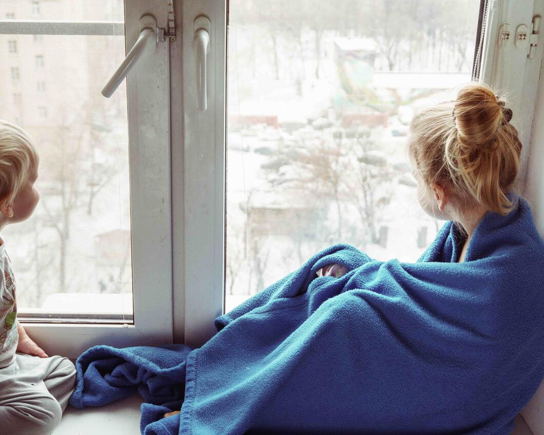 Twee kleine kinderen zitten voor een raam. Buiten is het koud. Het rechterkind heeft een blauwe deken om zich heen geslagen